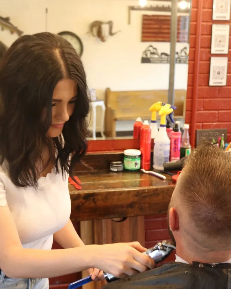 Barber at her station trimming a customer's hair near his beard
